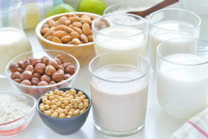 Glasses of various kinds of milk sit on a table next to bowls of nuts.