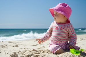 A baby sits in the sand at the beach and wears a pink hat and pink onesie that cover her whole body.