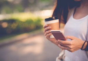 A woman holds a coffee in one hand and a cell phone in another. She uses her thumb to swipe across the phone's screen.