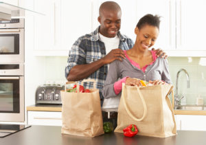 A woman stands in her kitchen and unloads her groceries. A man places his hands on her shoulders and smiles at the groceries.