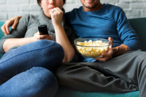 A woman and man sit together and watch a movie. They hold a bowl of popcorn.