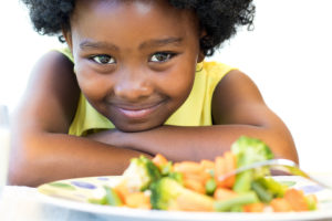 A young girl leans on a table and stares at a plate with broccoli and carrots. She smiles.
