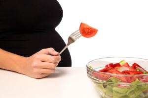 A pregnant woman sticks her fork into a bowl of salad and grabs a tomato.