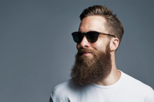 A bearded man wears pitch black sunglasses and smiles.