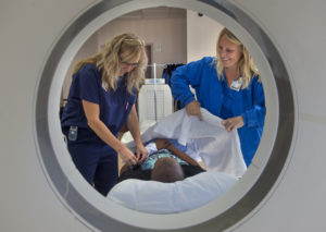 A young patient is being covered with a white blanket before he enters a CT scan machine. Two medical professionals are shown helping their patient.