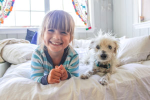 A young girl lies on a bed with a dog and smiles big.
