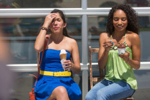 Two women sit together and eat ice cream. One of the girls holds her hand over her head. She appears to be having a brain freeze.