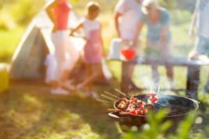A few people gather for a picnic. A little grill is in focus with kabobs on it.