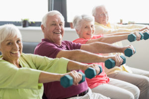 Five elderly adults work out together. They all hold their arms out and raise dumbbells.