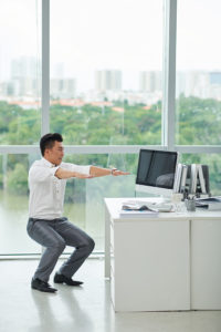 An adult squats at their desk. He is wearing business attire and looks at his computer screen while exercising.