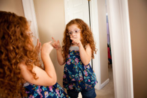 A young girl points at herself in the mirror and smiles.