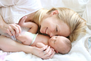 A woman lies in bed with her baby and smiles. The baby appears asleep.