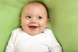 A baby smiles big and appears happy. The baby lies on a green blanket.