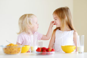 A young girl grabs a young girl's nose and smiles. They both sit at a table were strawberries, cereal and milk are shown.
