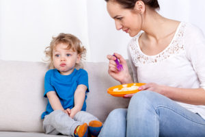 A woman holds a plate of food and tries to feed her kid food with a spoon.