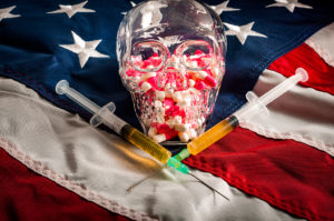 A glass skeleton head is filled with pink-and-white pills. The skeleton head sits on an American flag with two injection needles.