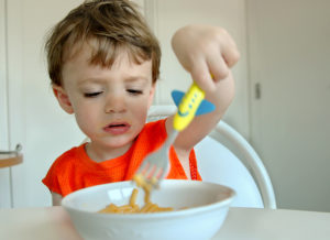 A toddler reaches into their bowl of mac and cheese with their fork and plays with it. The toddler looks unsatisfied.