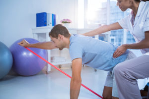 A man stands on his knees and pulls on a resistance band. A medical professional observes his exercises.