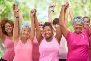 A group of women raise their hands together and smile. They all wear pink, breast cancer tank tops or T-shirts.