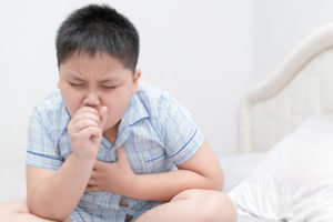 A young boy coughs into his hand while holding his chest.