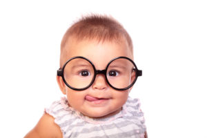 A baby girl smiles and licks her mouth. She wears big black glasses.