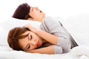 A woman covers her ears as she lies in bed with a man who appears to be snoring.
