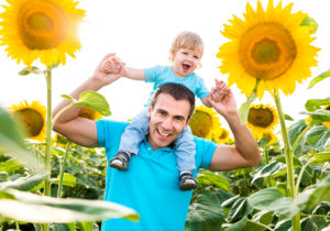 A man holds a toddler on his shoulders and smiles. They stand in a field of sunflowers.