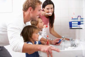 A mom and dad brushes their teeth with their two kids. They all hold a toothbrush as they stare into a mirror.