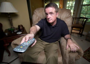 A man lounges in a chair at home and points a remote toward his TV.