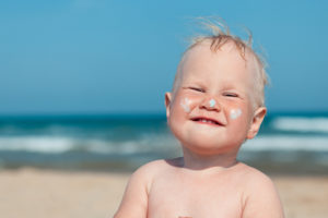 A baby wears sunscreen on his nose and cheeks. A beach is shown in the background.