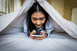 A young woman lies in bed and is on her phone. She hides under her sheets and blankets.