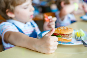 A young boy eats a hamburger and gives the camera a thumbs-up.