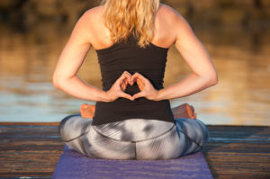 A woman sits on her yoga mat and places her hands behind her back. Her hands are shaped in a heart formation.
