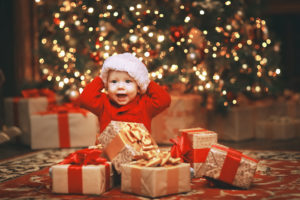 A baby wearing a Santa hat sits in front of a Christmas tree and smiles big. Multiple presents are placed in front of him.