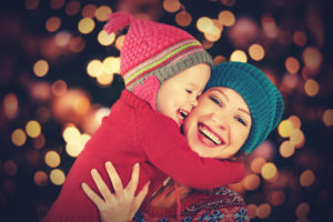 A woman holds a young boy in her arms and smiles big. The woman stands in front of a Christmas tree.
