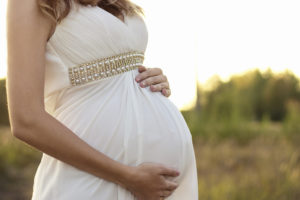 An expectant mother holds her pregnant belly. She wears a white dress and stands outside.