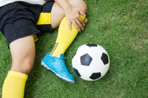 A soccer player lies in the grass and holds their knee. A soccer ball is shown next to the player.