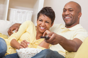 A man and woman sit on the couch and watch TV. The man holds the remote and smiles toward the screen, as the woman smiles big while grabbing popcorn out of a bowl.