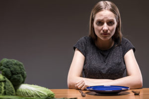 A young woman sits at a table with no food on her plate. She appears upset.