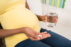 A pregnant woman holds a glass of water in one hand, and a vitamin D pill in the other.