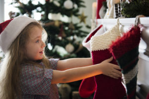 A young girl, wearing a Santa hat, reaches for a stocking on a fireplace.