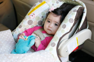 A toddler sits in a car seat inside a car and holds a stuffed animal.