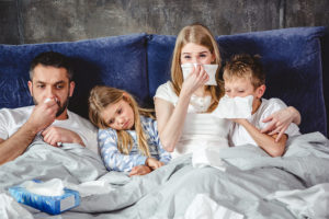 A family sits together in bed with tissues. They all appear sick.