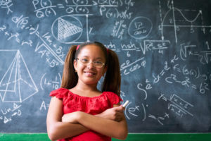 A young girl holds chalk next to a chalkboard full of math equations.