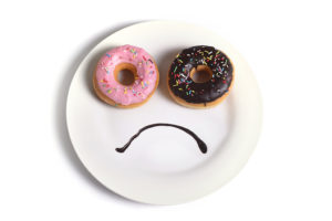 A white plate holds two donuts and chocolate syrup. The chocolate syrup forms a frowning face below the two donuts.
