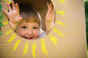 A little girl puts her hands through cardboard. The piece of cardboard has a circle cutout shaped like a sun.