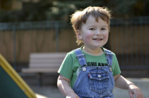 A little boy runs outside. He is wearing overalls.