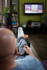 A bald man watches TV and lies on a couch with a remote in his hand.