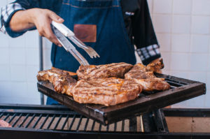A person wearing an apron uses tongs to grab a piece of steak off of a grill.