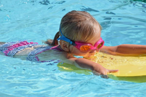 A little girl with pink goggles swims and holds onto a toddler swim float in a swimming pool.